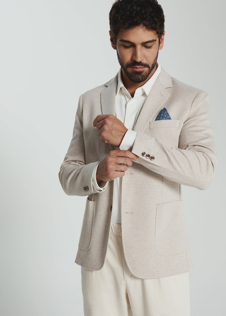 Man wearing a beige blazer and white shirt with a blue pocket square on a plain background